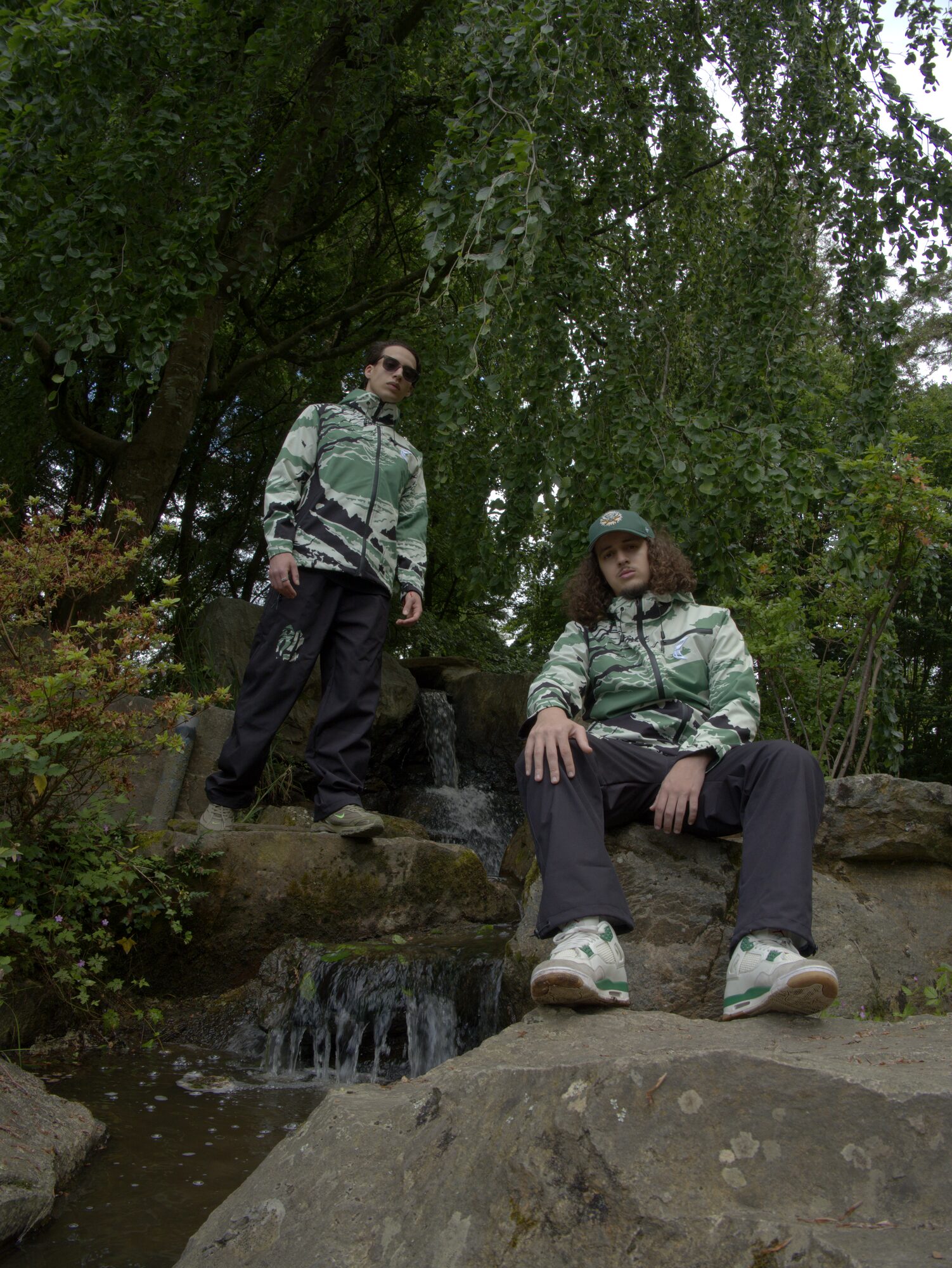 Deux hommes en vestes vertes sur des rochers près d'une cascade, vus en contre-plongée, projet 80 Cypper × Bluedrop Shop