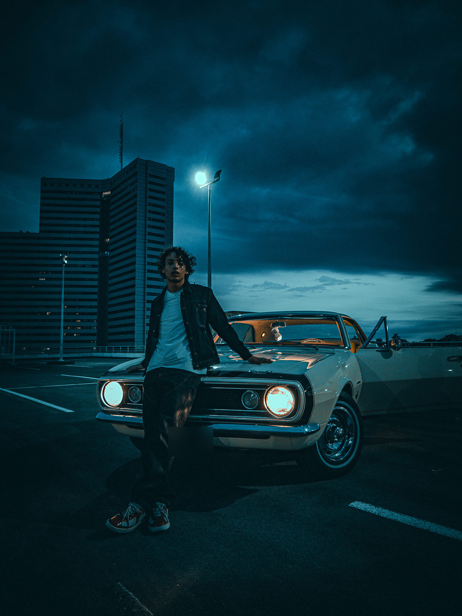 Homme en veste en jean appuyé sur le capot d'une voiture ancienne aux phares allumés, sur un parking de nuit, projet Feeling good