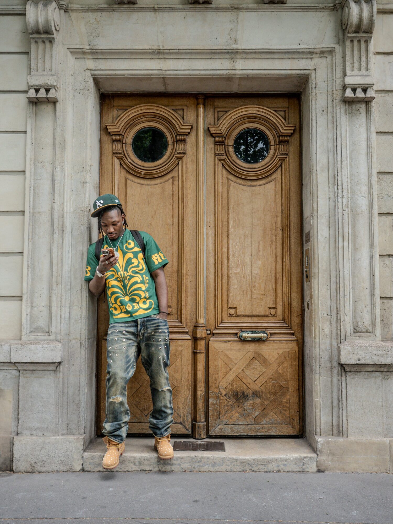 Homme en t-shirt vert et jaune et casquette A's, adossé à une porte en bois, shooting streetwear pour OG Shark