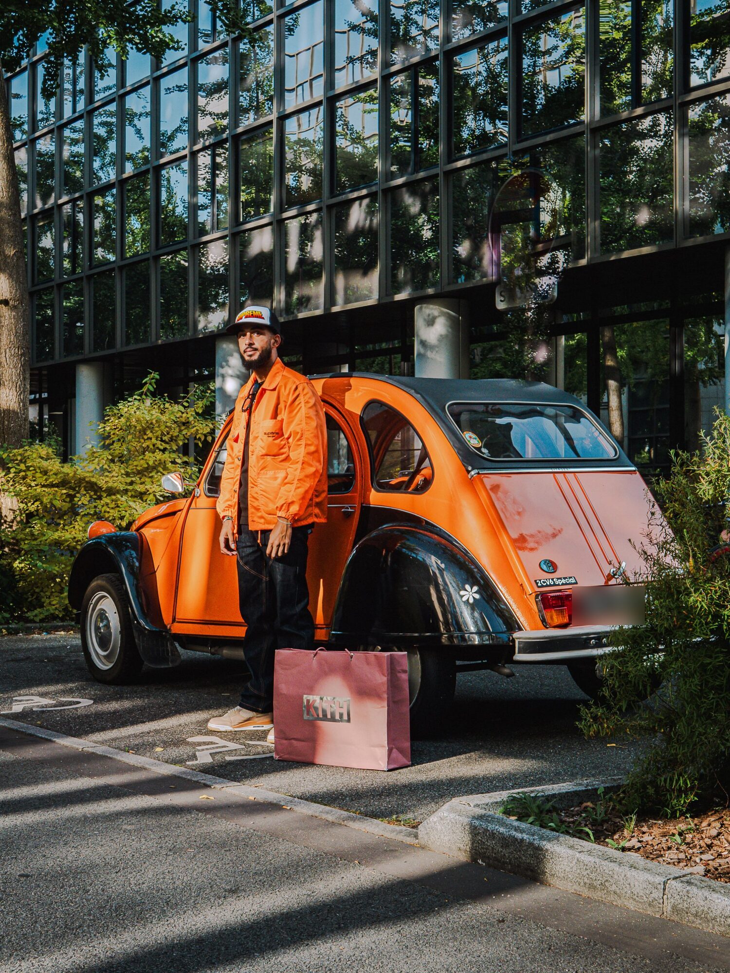 Homme en veste orange debout devant une 2CV orange, façade de verre en arrière-plan, projet personnel Orange